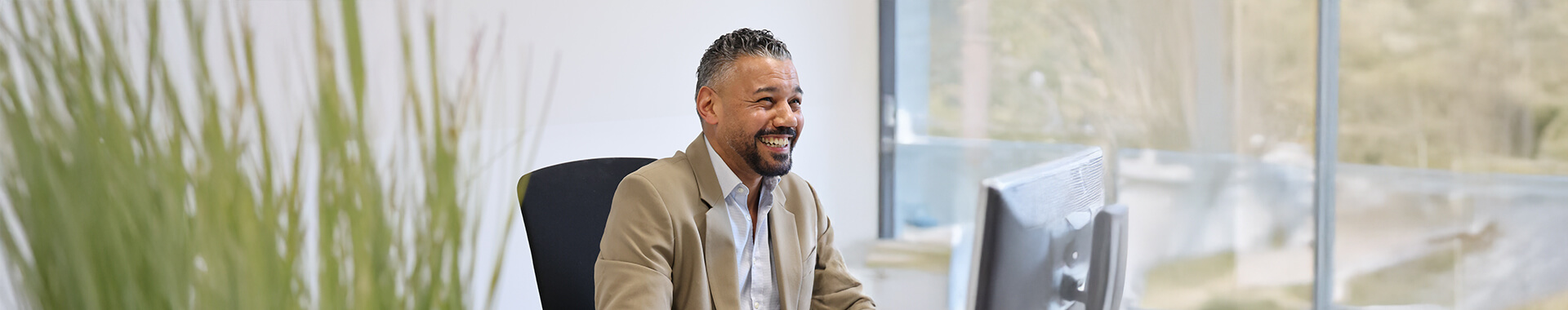 Freundlicher Geschäftsmann sitzt im Büro am Computer und lächelt, moderne Arbeitsumgebung mit Pflanzen